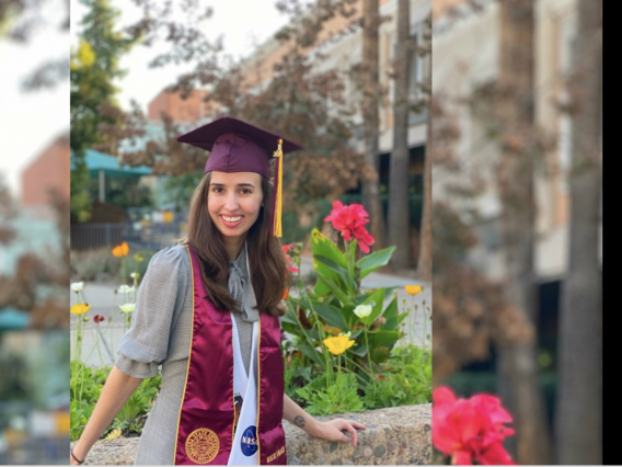 Elinor Saur in graduation cap and sash, posing outside by flowers.