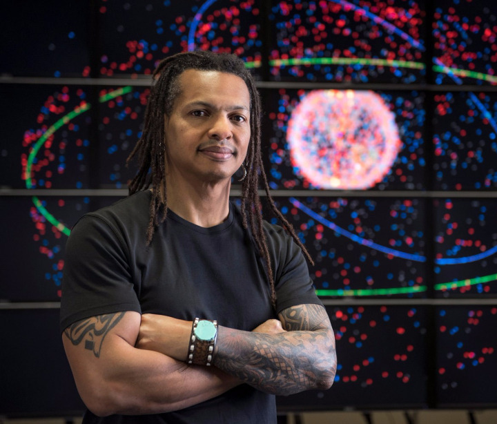 Moriba Jah stands in front of a multi-screen display of orbiting satellites.